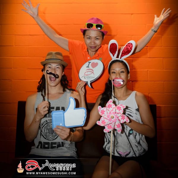Three people posing with fun props, including a mustache, bunny ears, and an "I love Awesome Sushi" sign.