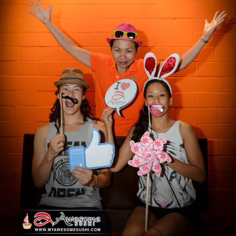Three people posing with fun props, including a mustache, bunny ears, and an "I love Awesome Sushi" sign.