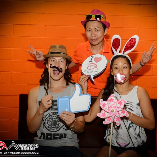 Three people posing with fun props like a mustache, bunny ears, and an "I love Awesome Sushi" sign.