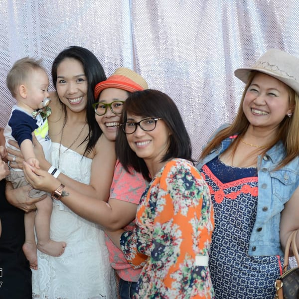 Four smiling women and a baby pose together against a shimmering silver backdrop.