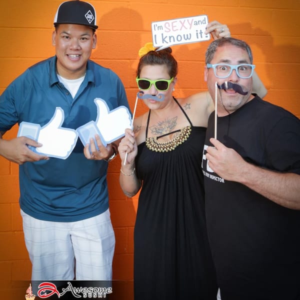 Three people posing with fun props like mustaches, sunglasses, and "thumbs up" signs against an orange wall.