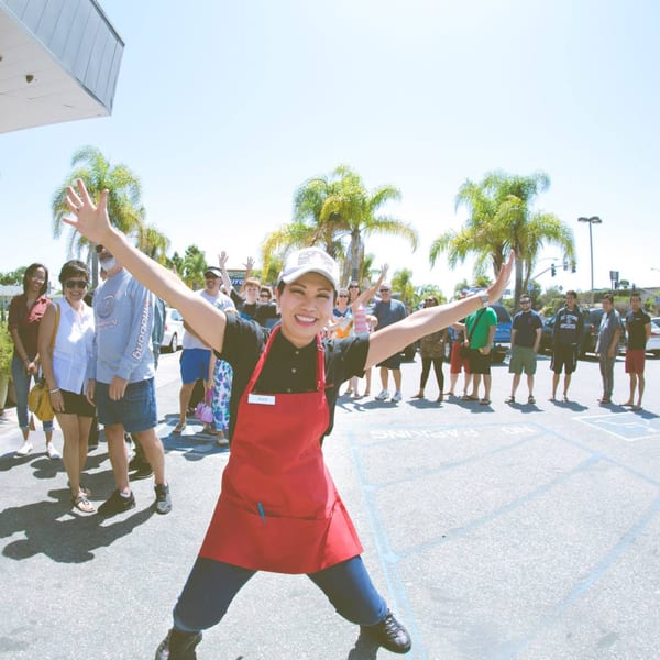 Awesome Sushi staff member in a red apron with arms outstretched, smiling at a crowd outdoors.