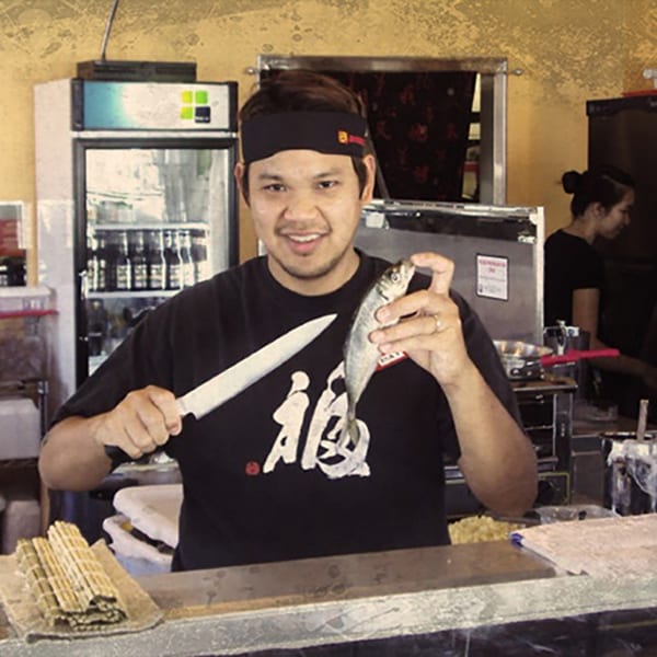 A smiling Awesome Sushi chef holds a knife and a whole fish, ready to prepare Japanese cuisine.