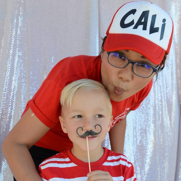 A woman in a red "Cali" hat and a boy with a fake mustache pose playfully.